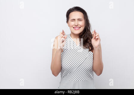 Portrait of hopeful beautiful young brunette woman with makeup and striped dress standing, clenched teeth, closed eyes and showing hope gesture. indoo Stock Photo