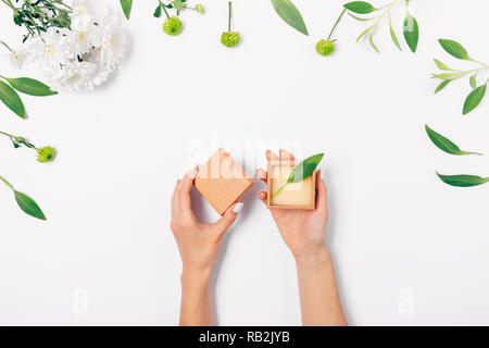 Woman's hands open small gift box of brown eco paper, flat layout of fresh flowers and green leaves on white background, top view. Stock Photo
