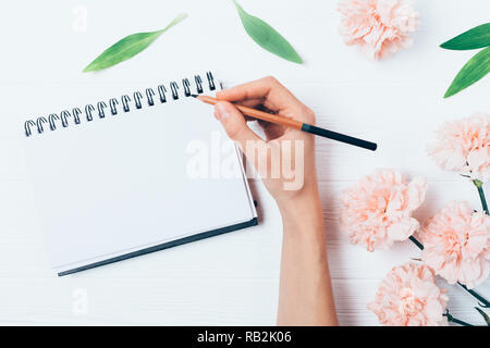 Top view of female hand holding pencil next to page of empty notebook among pink flowers and green leaves, flat layout on white wooden table. Stock Photo