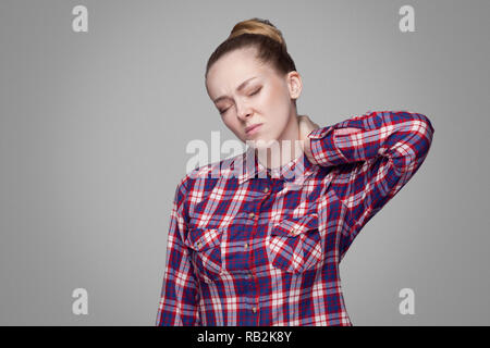 blonde girl in red, pink checkered shirt, collected bun hairstyle, makeup standing with closed eyes and touching her painful neck. indoor studio shot. Stock Photo