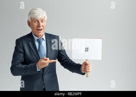 Senior business man studio standing isolated on gray wall holding white board pointing at number sign looking camera surprised Stock Photo