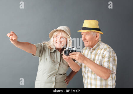 Senior couple tourists wearing beach hats studio standing isolated on gray wall holding camera looking for good shot smiling excited Stock Photo