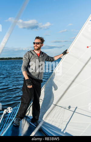 Young european man standing at edge of yacht looking at sea. Travelling on old boat with sail. Luxury lifestyle. Stock Photo