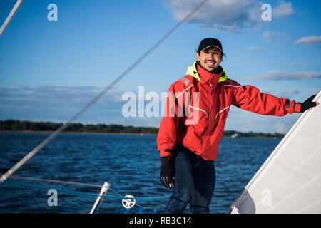 Young man skipper in red windbreaker and black cap sets the sails on a sailing yacht Stock Photo