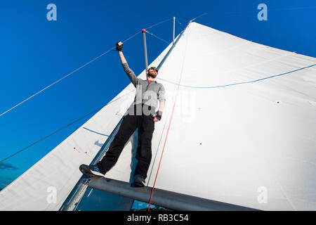 Nice and confident young man stands on yacht board and look at the sea through glasses. He is at the edge of yacht. Guy holding sail rope in his hand. Young man is happy and peaceful. Stock Photo