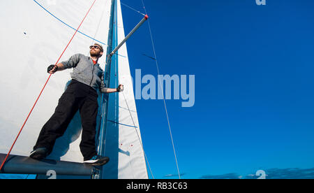 Yacht captain with a beard stands on sail boom on a sailing yacht, holding the rope in his hand and smiling, feeling happy. Adult yachtsman travelling around the world. Copy space Stock Photo