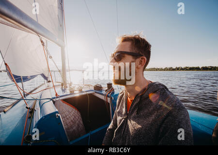 Man dressed in casual wear and sunglasses on a yacht. Happy adult bearded yachtsman close-up portrait. Handsome sailor on a boat smiling during regata on a sea or river. Stock Photo