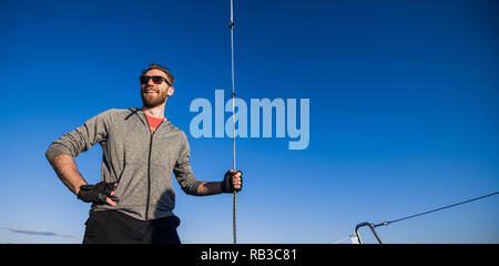 Young man wearing sunglasses standing on yacht stern and enjoying perfect autumn day under sails - sailing holidays concept Stock Photo