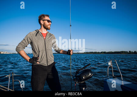 Young man on a catamaran standing and holding rope in front of a blue sky during cruising. Stock Photo