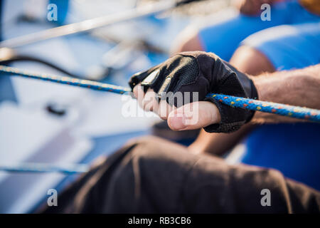Close up of sailor winds aroung rope by using handle for coiling. He works with both hands. Young man stands on yacht. He is strong Stock Photo