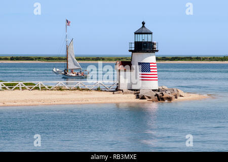 Brant Point lighthouse, with its American flag wrapped around its wooden tower, guides a sailboat out of Nantucket Island harbor in Massachusetts. Stock Photo