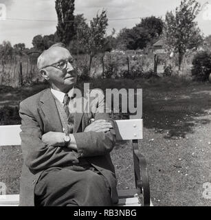 1964, an elderly man wearing a jacket and tie sitting on a wooden bench in a park outside enjoying the sunshine and fresh air, England, UK Stock Photo