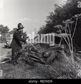1964, a fireman using a hose to put water onto a burnt-out car on the  grassy verge of a country road, England, UK. Stock Photo