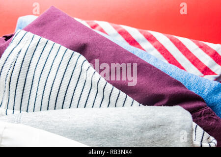 Pile of different clothes on red background. Top view of variety of shirts and jeans laying one on another. Stock Photo