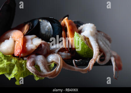 Mixed raw delicious seafood with lime slices on a dark background. Small octopuses, shrimps and mussels on a leaf of salad. Mediterranean kitchen, hea Stock Photo
