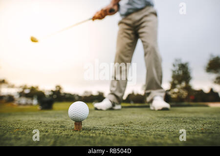 Golf ball on tee with man blurred at back about to make a shot at golf course. Focus on golf ball on tee with male golfer taking a shot. Stock Photo
