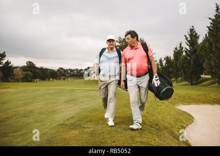 Two senior friends walking through the lawn with golf equipment and talking. Senior golf players walking together on the golf course. Stock Photo
