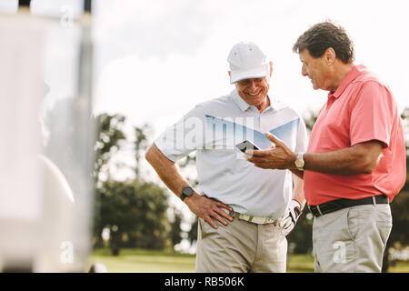 Two golf player standing together using mobile phone. Senior golfers looking the scores on phone after the game on the golf course. Stock Photo