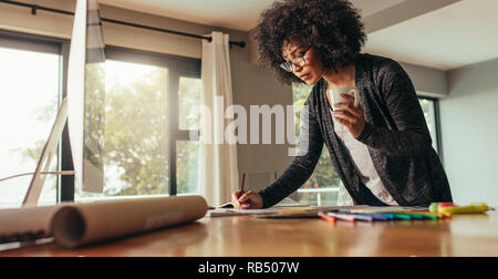 Architect working on plans and blueprints. Woman with cup of coffee making notes in a book at home office. Stock Photo