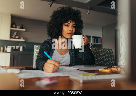 African woman working at her desk making notes looking at computer monitor and drinking coffee. Female architect working at her home office desk. Stock Photo