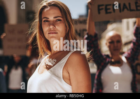Woman activist with word strong written on her body standing outdoors. Strong young female activist protesting outdoors with her group in background. Stock Photo