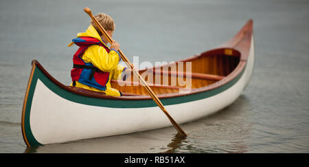 Young boy wearing a yellow raincoat and a life jacket tries to paddle a wooden canoe. Stock Photo