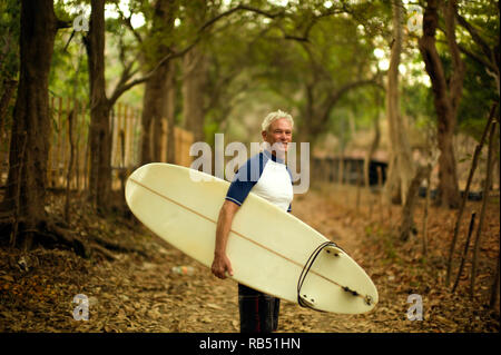 Mature man smiles as he stands on a rural path carrying a surfboard and poses for a portrait. Stock Photo