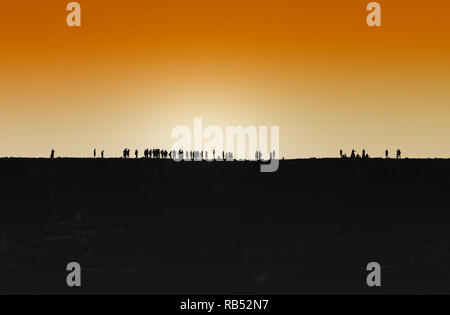 Group of people socialising and enjoying themselves on an elevated ridge outdoors, silhouetted against an orange sky at sunset. Stock Photo