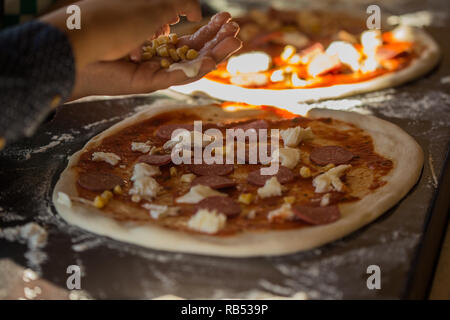 Pizza baking process. Child's hands putting corn on pizza with mozarella and salami Stock Photo