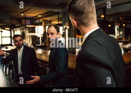 Three young men in suits lok at each other. One of them stand with back to camera. Office workers stand in pub. Stock Photo