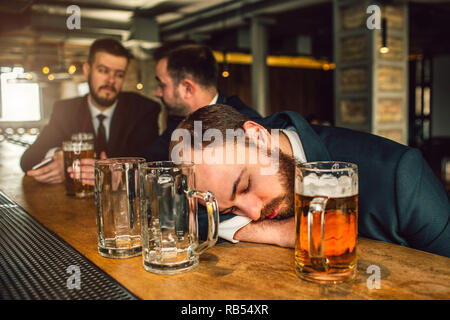 Tired young man in suit sleep on bar counter. He is drunk. there are two empty mugs and one full with beer. Other two young men sit behind. Stock Photo