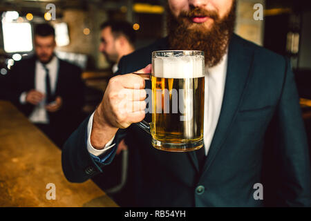 Cut view of bearded man in suit hold beer mug. Other two office workers sit behind. Stock Photo