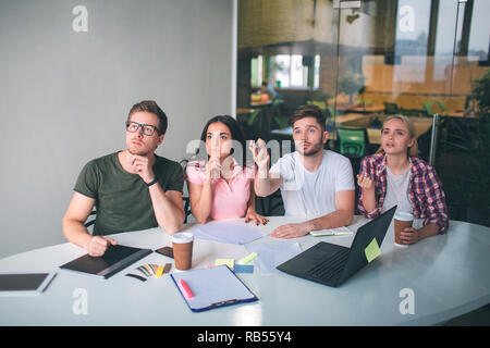 Serious and concentrated team members sit at table and look up. They work. Guy on right pointing. People work together on white room. Stock Photo