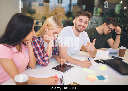 Handsome young bearded man sit among his coworkers and hold big thumb up. He smiles on camera. Other people work and look down. They all are in one room. Stock Photo