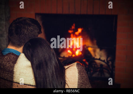 Brunette young man and woman sit together and look at fireplace. She hold her head on his shoulder. They covered with blanket. Stock Photo