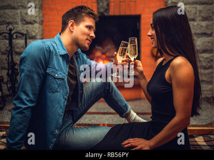 Serious young man sit on floor with his girlfriend and look at her. She smiles to him. They hold glasses with champaigne. Couple sit at fireplace. Stock Photo