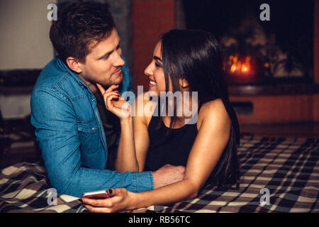 Young man and woman lying on floor together. She look at him. Young woman hold hand under man's chin. They are not far from fireplace. Stock Photo