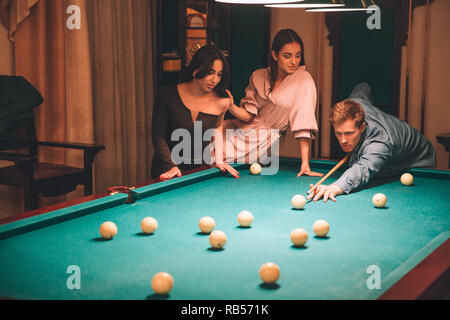 Concentrated young redhead man aiming into billiard ball. He look straight forward. Young woman in pink dress sit at billiard table. Asian brunette st Stock Photo