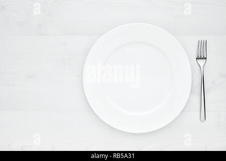 Empty white round ceramic plate and fork on white wooden table with copy space Stock Photo