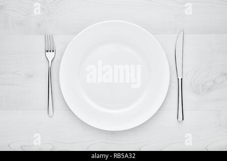Empty white round ceramic plate, fork and knife on white wooden table with copy space Stock Photo