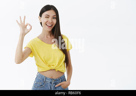 Good-looking female fitness trainer assuring she will help winking joyfully showing okay or excellent gesture with raised fingers holding hand in pockets standing confident, delighted over grey wall Stock Photo