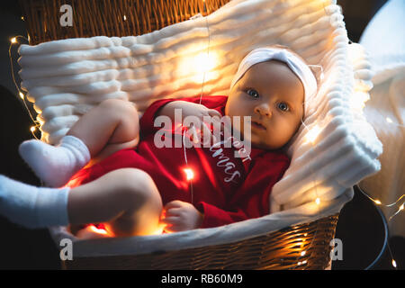 Lovely, beautiful newborn girl lying in basket in a white hat, new year and Christmas decorations, garland on the wall, with a flashlight. card, text. Stock Photo