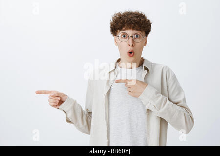 Waist-up shot of amazed young male friend with round glasses and curly haircut widen eyes folding lips with wow sound pointing left excited and astonished as if witnessing incredible thing happened Stock Photo
