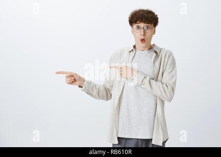 Impressed and curious attractive young funny guy with curly hair in round eyewear folding lips in amazement and widen eyes at camera pointing left with index fingers to indicate fantastic copy space Stock Photo