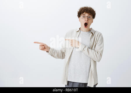 Omg no one ever believe. Portrait of impressed and astonished excited funny young guy with curly hairstyle in round prescribed glasses dropping jaw yelling wow as pointing left at awesome product Stock Photo