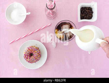 Hand pouring milk into a glass filled with ice cubes and cold brew coffee and a pink frosted donut with colorful sprinkles on a plate on pink backgrou Stock Photo