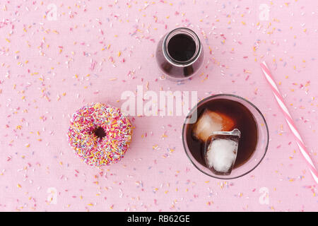 Pink frosted donut with colorful sprinkles, a glass filled with cold brew coffee, cold brew coffee concentrate in a milk bottle and a straw on pink ba Stock Photo