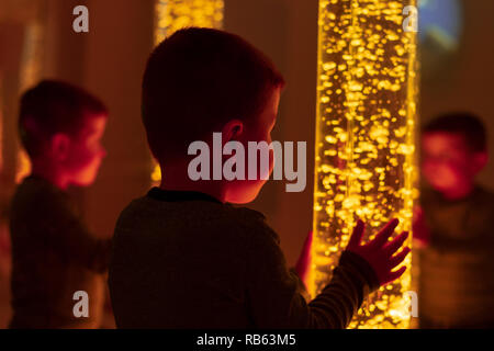 Child in therapy sensory stimulating room, snoezelen. Child interacting with colored lights bubble tube lamp during therapy session. Stock Photo