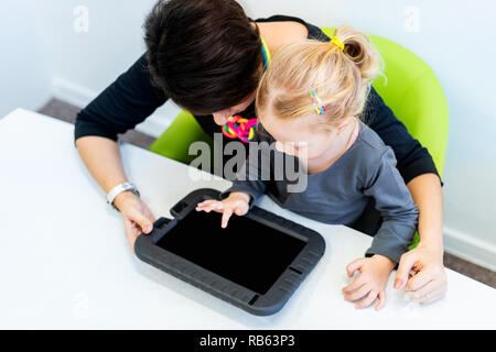 Toddler girl in child occupational therapy session doing sensory playful exercises with her therapist using digital tablet. Top view. Stock Photo