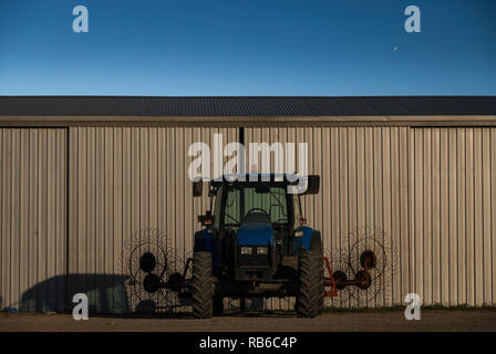 A night photograph of a tractor and rake against a corrugated shed with dusk sky in background and star trails in the sky Stock Photo